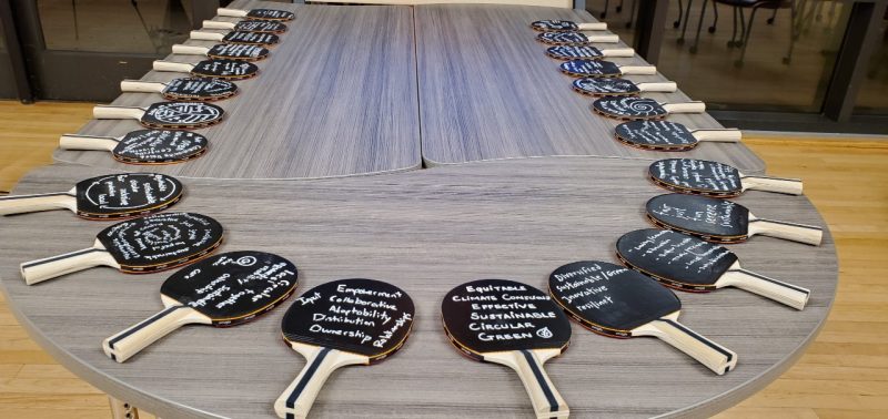 This photo shows a table with a couple dozen ping pong bats arranged around the perimeter. Each bat has a black surface with white writing on it.