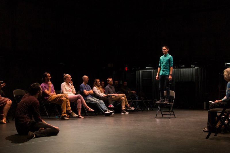 This photo shows a young man seated on the floor and another standing on a chair. They are looking at each other. Seated audience members look into the performance space from two sides.