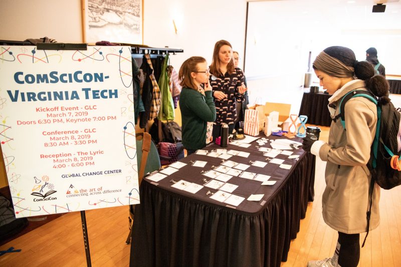 This photo shows a young African American woman in a coat and headscarf and wearing a backpack. She is looking at a conference registration table covered with nametags. Two white women stand behind the table. A sign on an easel reads "ComSciCon-Virginia Tech."