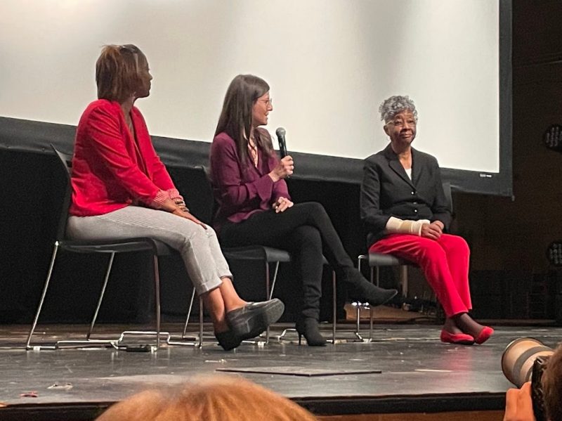 Three individuals sit on a stage with a white screen behind them, a White woman sitting between two Black women. The White woman holds the microphone while the left woman watches her. The woman on the far right looks out at the crowd.