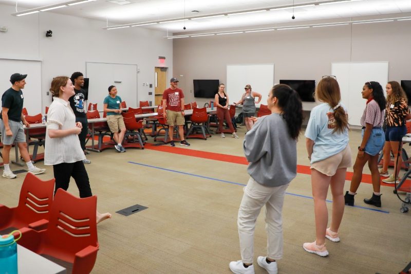 A group of students gather in a classroom. A thick red line and a much thinner blue line decorate the floor. The groups of people stand on opposite sides of the room.
