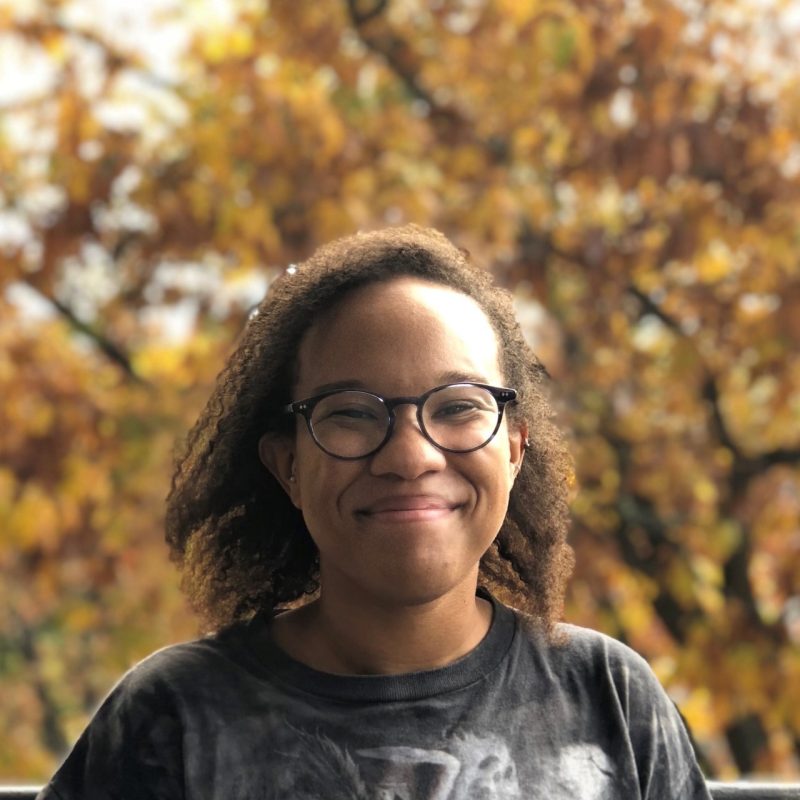A young African-American woman wearing a dark grey t-shirt and black glasses with colorful autumn leaves in the background.