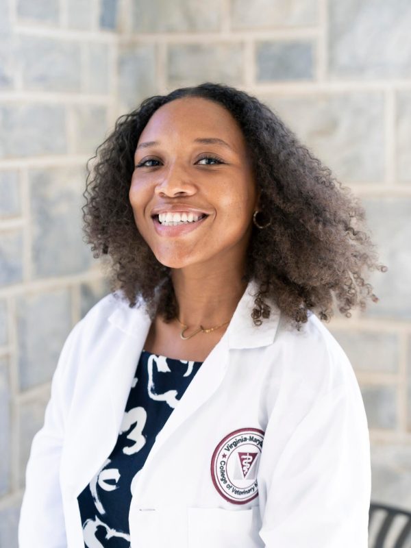 A young Black woman with shoulder-length hair is set against a gray brick background. She wears a blue and white patterned dress below a white lab coat with the Virginia-Maryland College of Veterinary Medicine logo on the left breast.