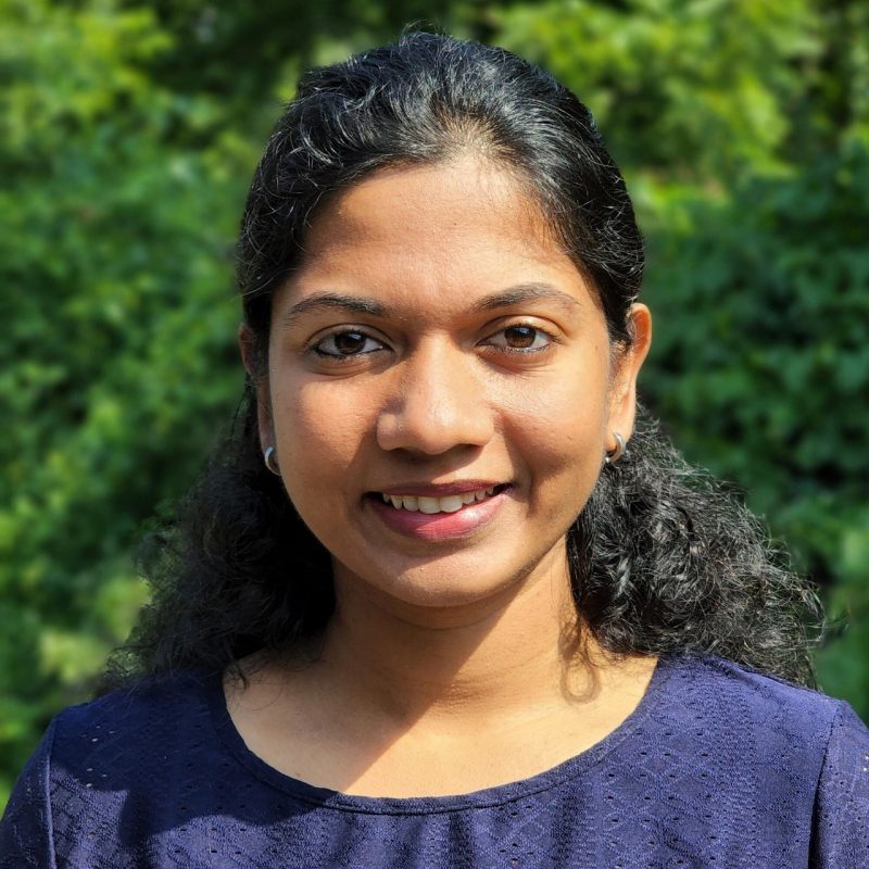 A young Indian woman with long dark, curly hair tied back into a half-up hairstyle. She is wearing a blue shirt and has trees in the background in professional headshot fashion.