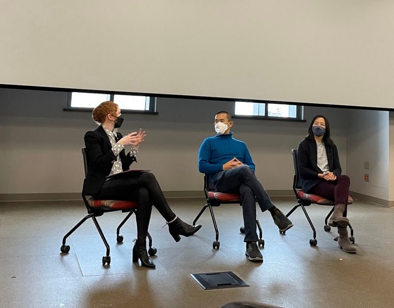 Ed Yong, an Asian man wearing a long-sleeve blue shirt,  sits between two women wearing dark pants, dark blazers, and light shirts. The woman on the right with red hair has her hand held out as she explains something.