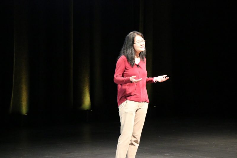 A woman stands on stage looking out into the audience and gesturing with a shrug