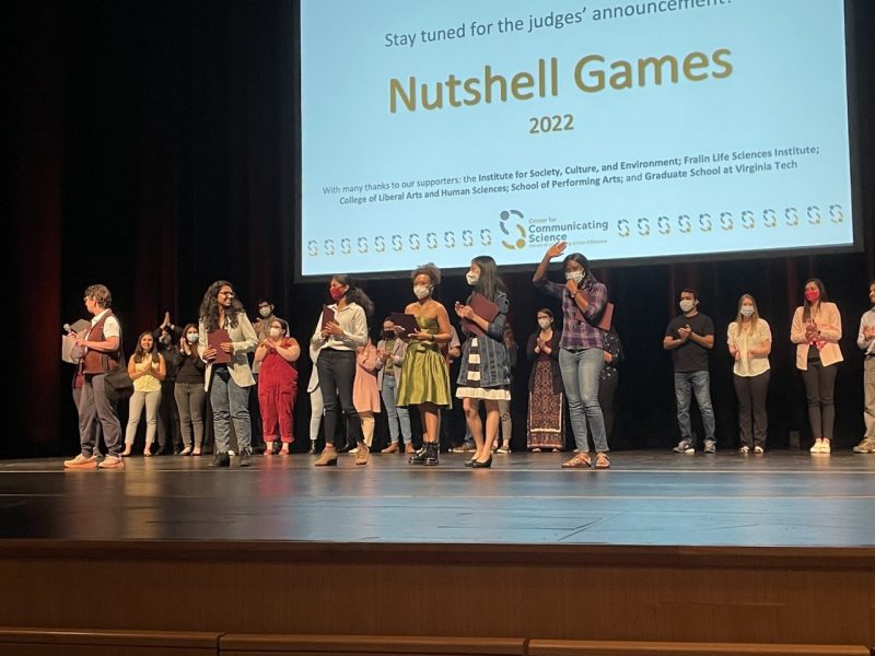 This photo shows a group of young adults lined up on a stage with a large screen behind them that reads "Stay tuned for the judges' announce. . . Nutshell Games" and has the logo for Virginia Tech's Center for Communicating Science.