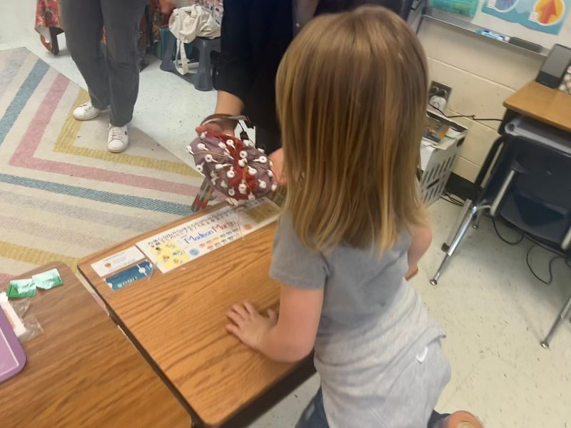 A little girl kneels on her chair at her desk, lightly touching an EEG cap being displayed to her. by a woman standing on a colorful, striped rug