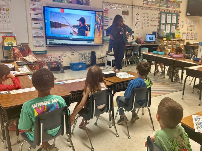 Woman standing at the head of the kindergarten class with focused students looking at her and screen with a man fishing.