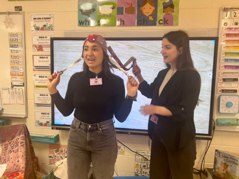 One woman stands at the front of a kindergarten classroom, eyes wide and smiling, donning an EEG cap, displaying the cap's straps while another smiling woman displays straps at the back of the cap.