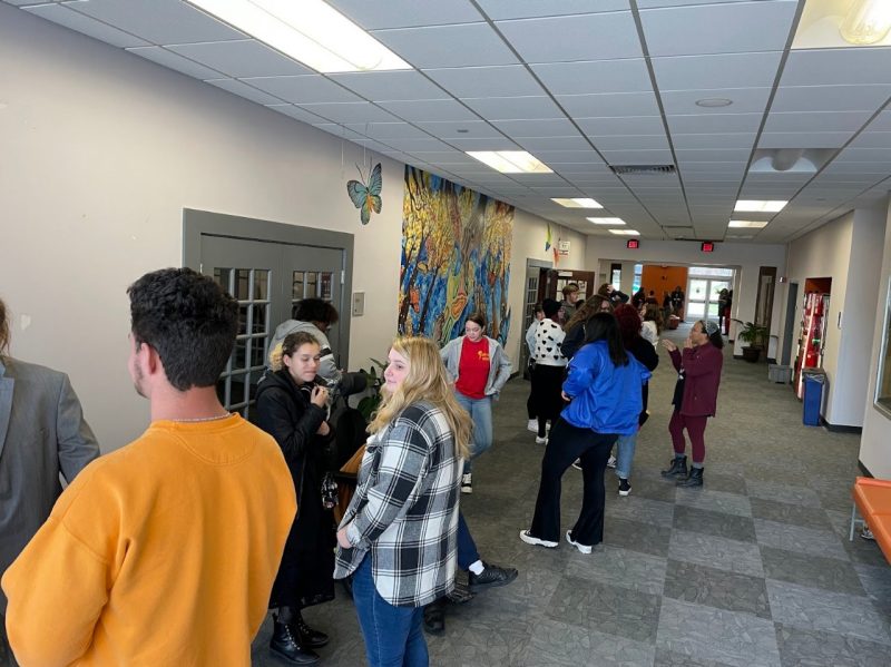 A hallway with gray checkered floor and white walls is filled with students including a man with dark hair and a bright yellow sweatshirt and a woman with blonde hair and a black and white flannel shirt.