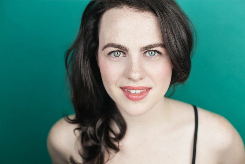 A young white woman with dark brown hair and bright blue eyes against a turquoise background.