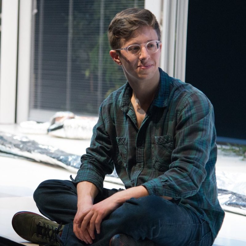 A young man with light brown hair in jeans, glasses, and a blue flannel shirt.