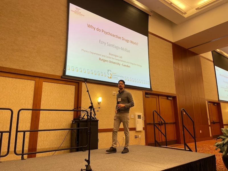 This photo shows a young white male wearing khaki pants and a gray sweater walking toward the camera on a stage. Behind him is a large screen projecting a slide the says Why do Psychoactive Drugs Work? and his name Ezry Santiago-McRae, along with a logo for the Virginia Tech Center for Communicating Science.