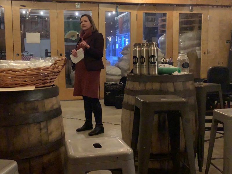 A woman wearing a red dress, black sweater, and dark tights points toward the crowd at the Rising Silo Brewery. She also holds a paper in her hand. Around her are empty water bottles and a basket of plastic bags.