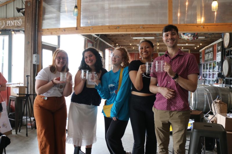 Five young adults (three white women, one Black woman, and one man) stand side by side smiling at the camera and holding up clear glass beakers with handles.