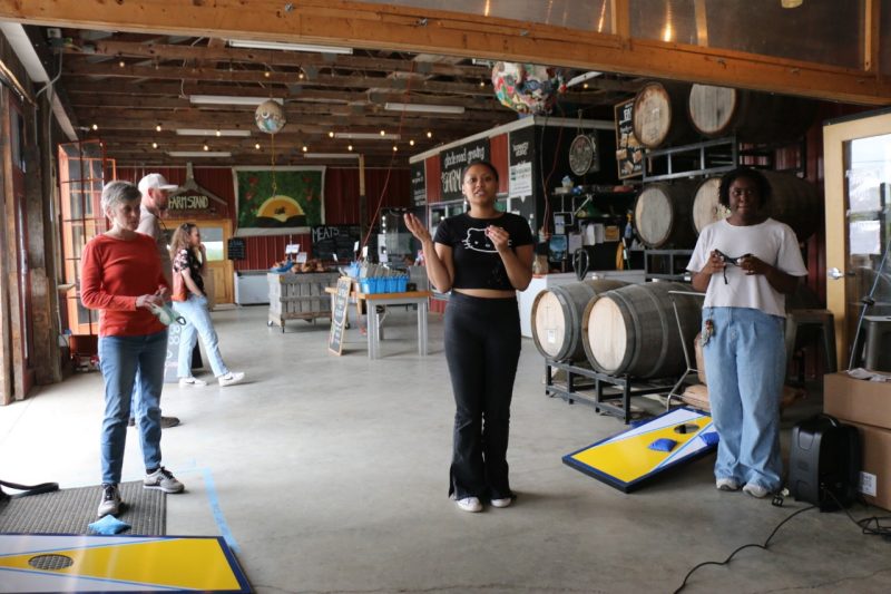 Alana Hutchinson talks in the middle of two participants, each holding goggles and standing next to cornhole goals.