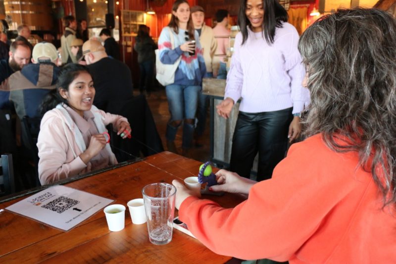 One participant puppets an arm, which is holding a ball, while another participant steadies the arm and cup on the table.