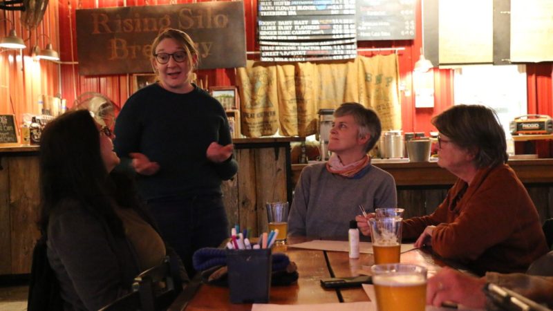 Maria DeNunzio stands, speaking with three participants seated around a table.