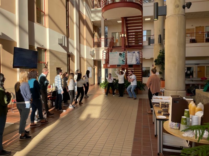 A building’s hallway is lined with people on the left side. In the center of the image, three people wearing white pose, the two on the outside crouching with their hands clasped, and the woman in the middle forming an O with her hands. A Panera breakfast spread lines the right side of the hallway.