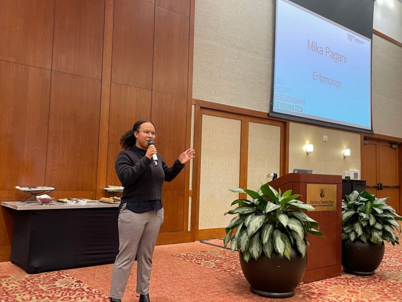 This photo shows a young Black woman speaking into a microphone. Behind and to her left is a large projection screen with a slide reading "Mika Pagani" and "Entomology."