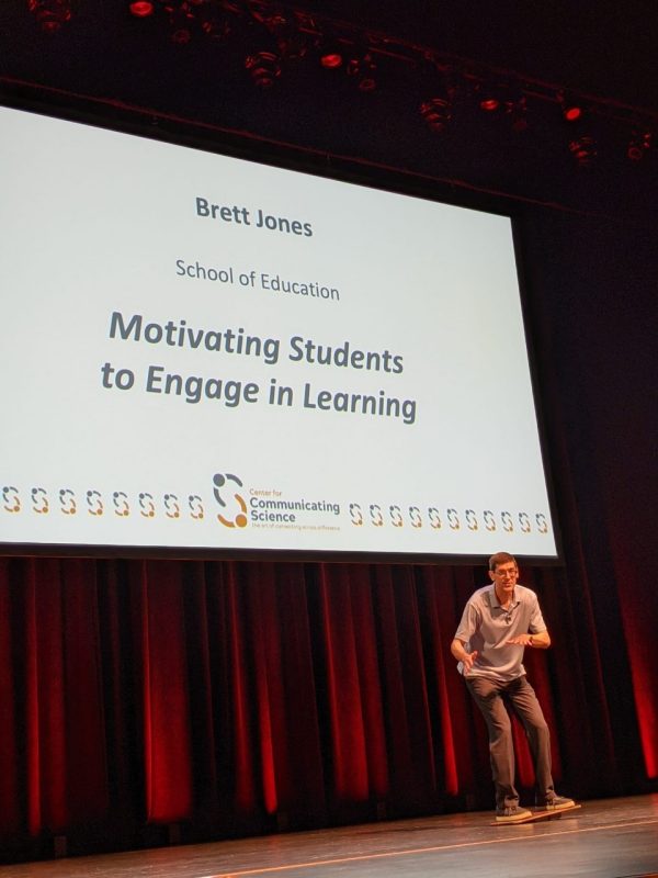 This photo shows a white man trying to keep his balance on a balance board. He is standing on a stage with red curtains as a backdrop. Behind and above him is a slide reading "Brett Jones, School of Education, Motivating Students to Engage in Learning." 