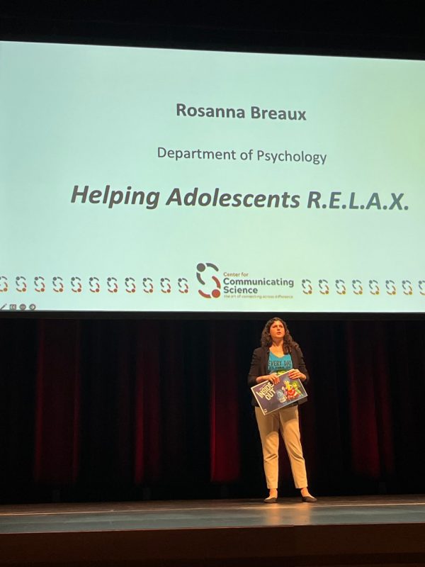This photo shows a woman holding a sign or placard, standing on stage, and behind her is a slide that reads "Rosanna Breaux, Department of Psychology, Helping Adolescents R.E.L.A.X."