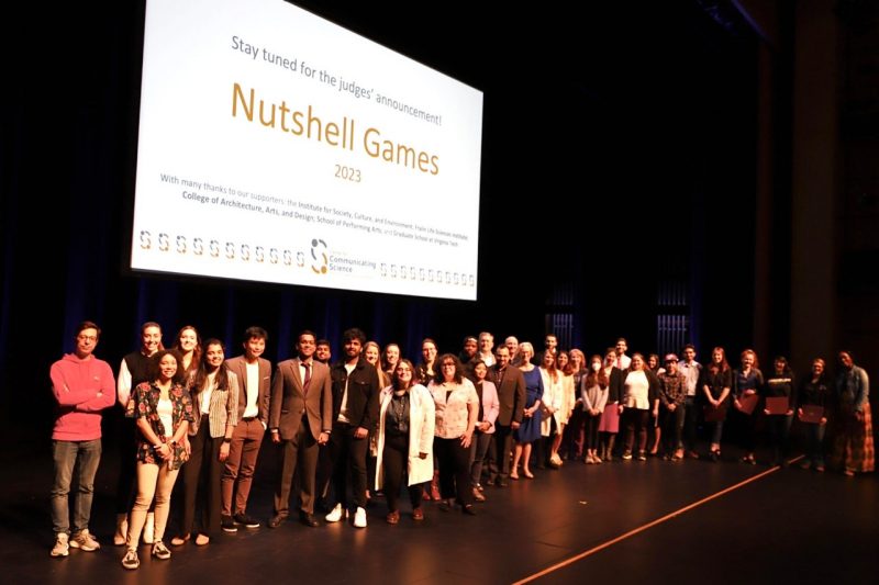 This photo shows a group of approximately 40 people lined up across a stage and in front of/below a projected slide that reads "Stay tuned for the judges' announcement! Nutshell Games 2023"