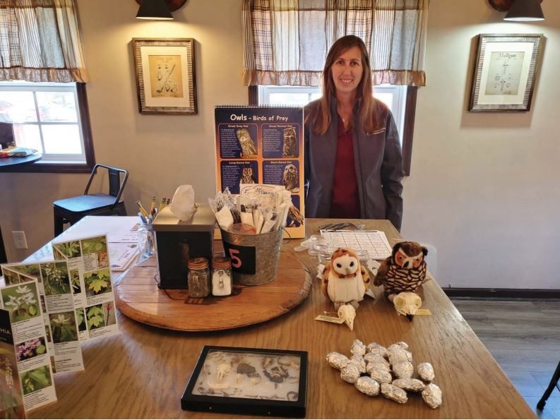 This photo shows a young white woman standing behind a table that has two stuffed animal owls on it along with a foldout tree identification  guide, some small foil-wrapped packages, and a framed class case with skeletal remains in in.