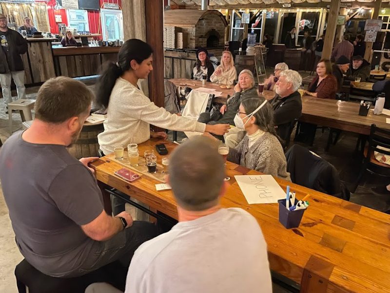 This photo shows a young Indian woman in a white sweater holding a small mic to the masked face of a middle aged white woman. The backs of two men are in the foreground and other people can be seen in the background. The scene is interior at a brewery.