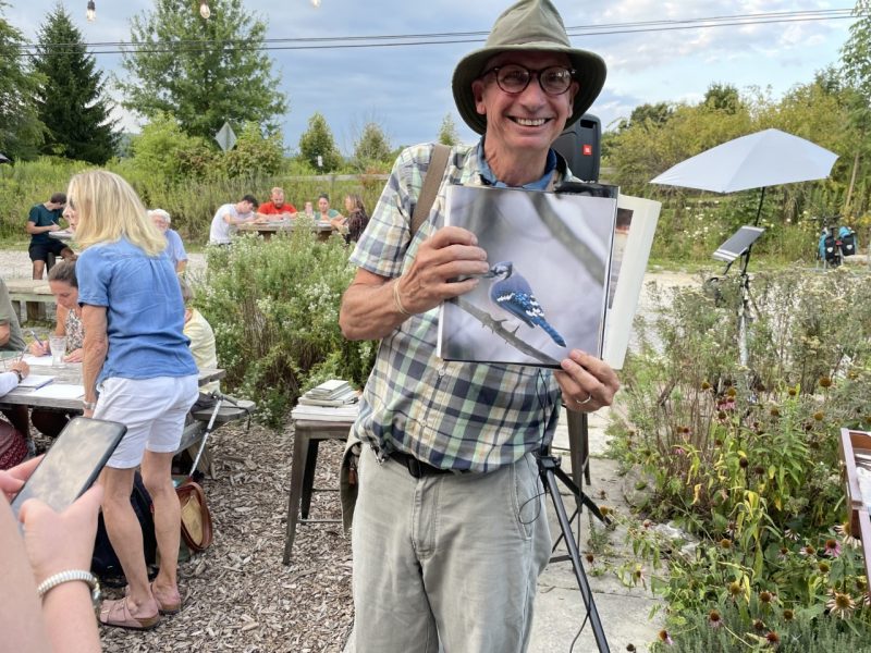 This photo shows a man wearing a plaid shirt, khaki pants, a khaki hat, glasses, and a big smile holding a book with a photo of a blue jay.