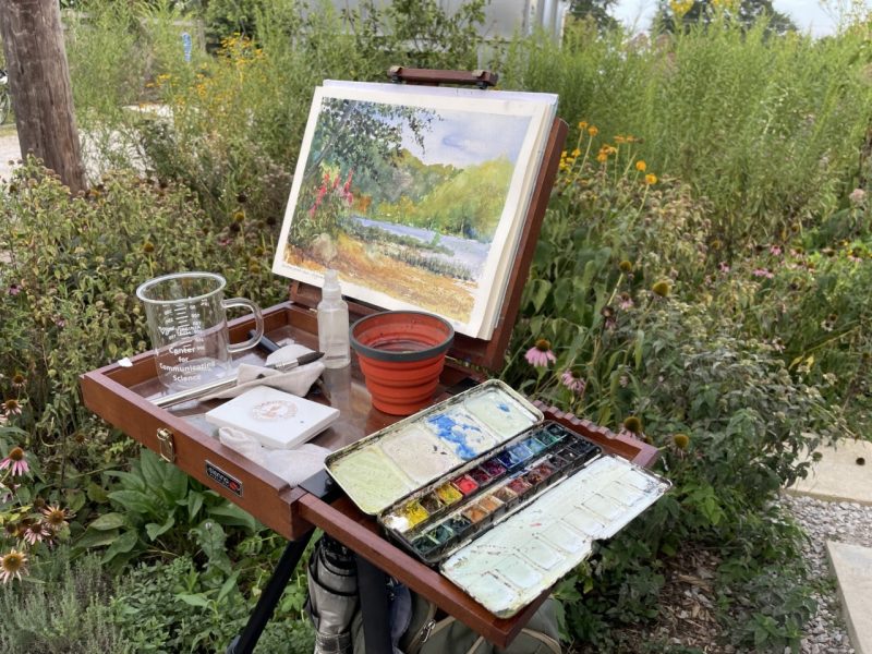 This photo shows an easel and art supply box. On the easel is a watercolor painting of a river scene.