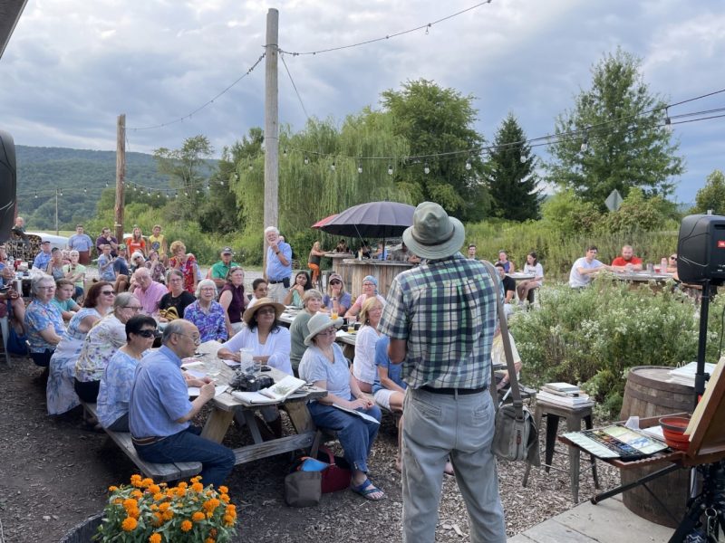This image shows the back of a person speaking to a crowd of people seated at picnic tables in an outdoor venue.