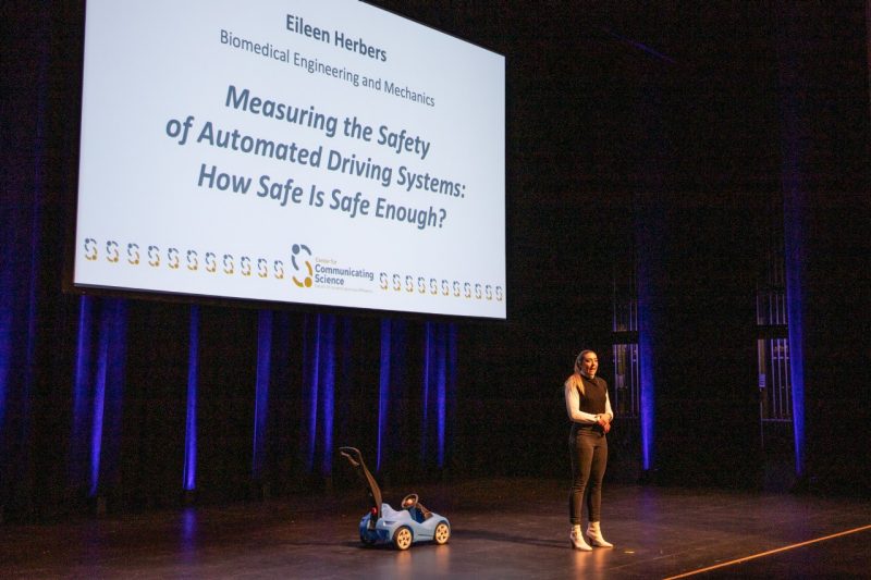This photo shows a young white woman standing on a stage with a toddler's blue plastic push car behind her. Also behind her is a slide that reads "Eileen Herbers, Biomedial Engineering and Mechanics, Measuring the Safety of Automated Driving Systems: How Safe is Safe Enough?"