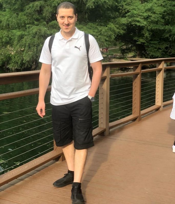 Abdu Alkishe A photo of a man on a bridge with a lake in the background. He is wearing a white colored puma shirt, a black shirt and pants, and his right hand is in his pocket with a watch.
