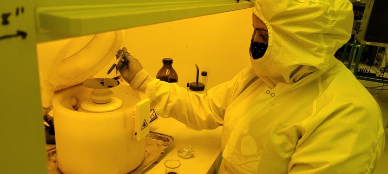 This photo shows a woman from the chest up clad in a white lab garment that covers all but her nose and eyes. She is also wearing a black face mask, so only her eyes, eyebrows, and a bit of forehead are visible. She is in a laboratory and is working with an instrument that resembles a small potter's wheel inside a fume hood.