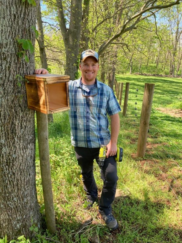 This photo shows a young white male dressed in dark pants, a short-sleeved blue plaid shirt, and a baseball cap, smiling at the camera. He is standing next to a wire fence with woods to the left of the photo and pasture to the right. He is holding a power drill and has his other hand on a wooden box (about 12"x12"x12") on a 5-foot post.
