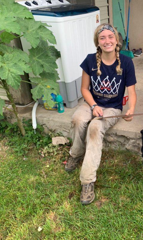 This photo shows a young white woman with blond braids seated on a concrete stoop and holding a machete blade in her lap as she smiles at the camera.