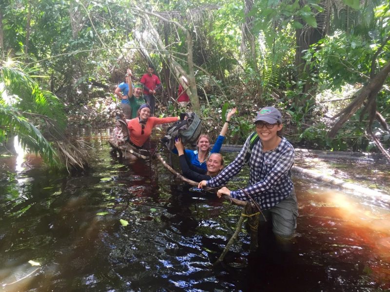This photo shows 8 people thigh- to chest-deep in water and holding onto a rope or branch as they cross a river in a rainforest.