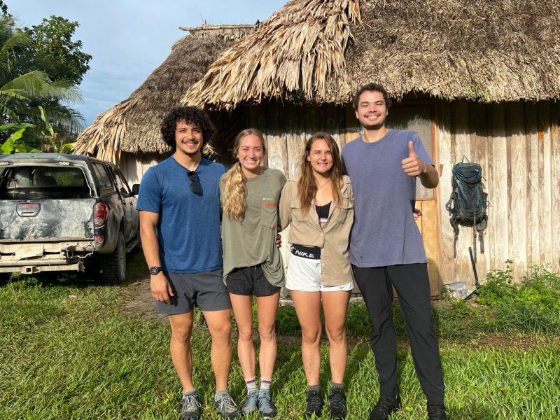 This photo shows four young people lined up for a photo in front of a thatched building. In the background is an old station wagon and another thatched wooden building.