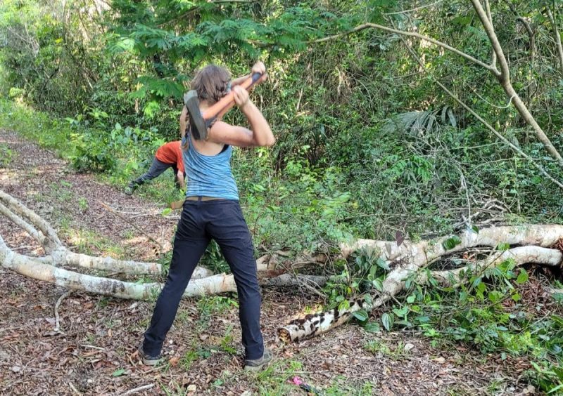 This photo shows a person from the back with a large axe over her shoulder, poised to swing down into a fallen tree blocking a road.