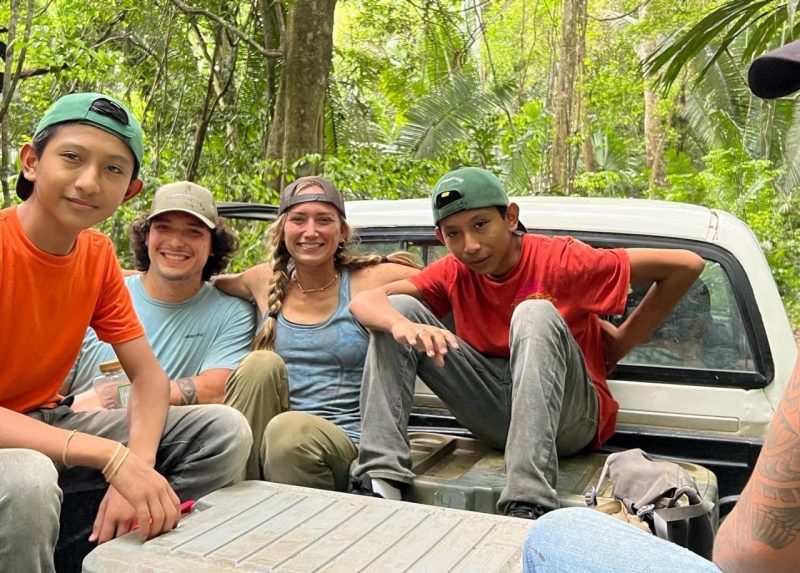 This photo shows four young people in the back of a pickup truck seated around a picnic cooler. Surrounding them is lush green forest.