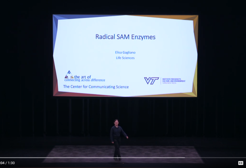 This clip from a video shows a young Caucasian woman with dark hair dressed in black slacks and a black top. She is walking out into the middle of a large stage with a projected image behind her that reads "Radical SAM Enzymes/Elisa Gagliano/Life Sciences/The Center for Communicating Science/the art of connecting across difference/VT/Institute for Society, Culture, and Environment."