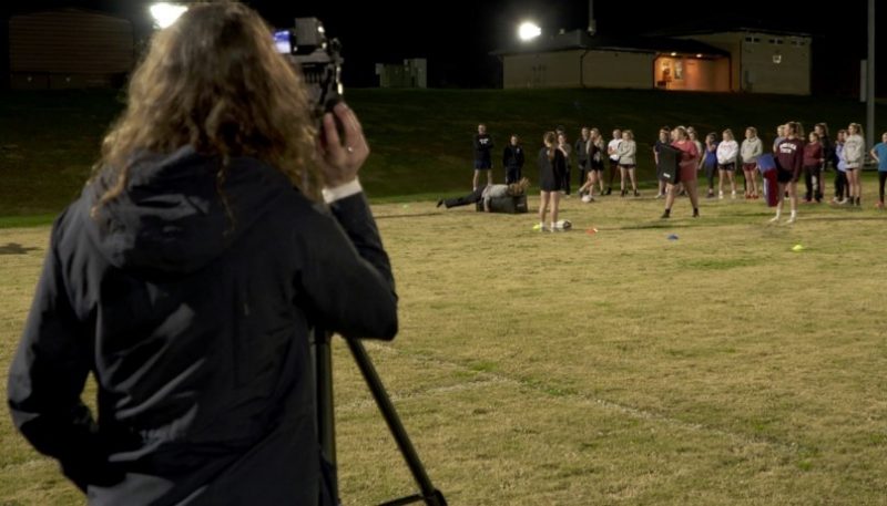 This photo shows the back of a person with long brown hair wearing a black jacket and standing behind a camera on a tripod. In the background are players of a team support.