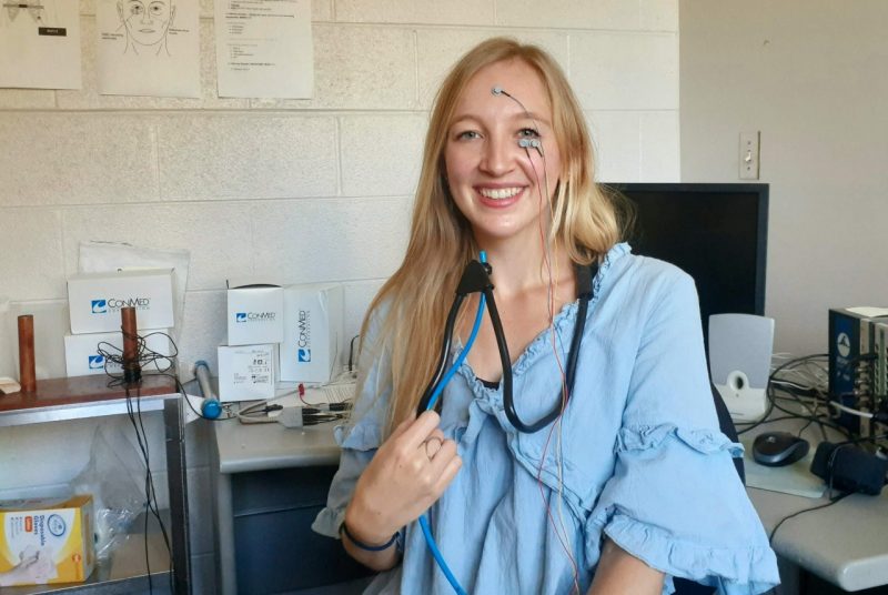This photo shows a young white woman with long blond hair clad in a blue blouse and smiling at the camera. She has wires and electrodes taped to her forehead and cheek and a black apparatus draped over her shoulders and around her neck.