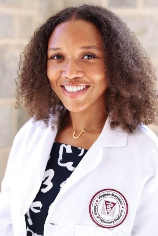 This photo shows the head and shoulders of a young Black woman with light brown skin and dark brown shoulder length curly hair, smiling directly at the camera and wearing a white lab coat.