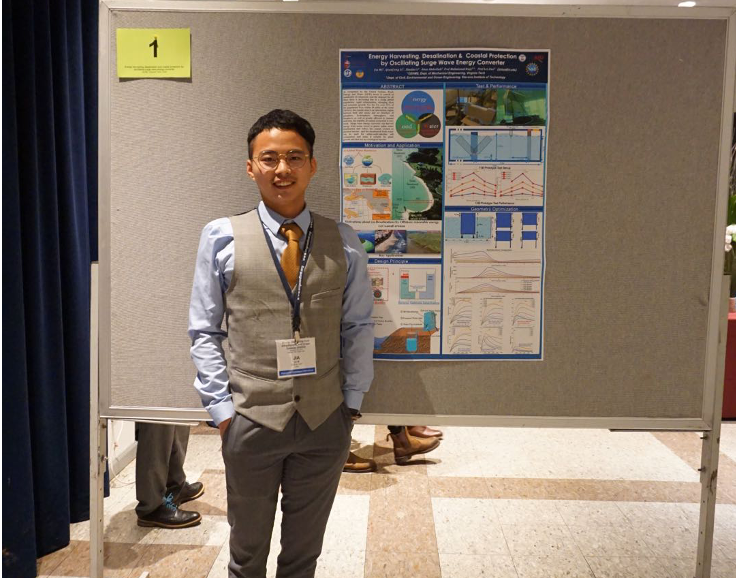 This photo shows a young Asian man with short dark hair clad in a light blue shirt, grey vest, grey trousers, and brown tie and shoes standing in front of a colorful research poster. He has a nametag around his neck and a cheerful smile on his face.