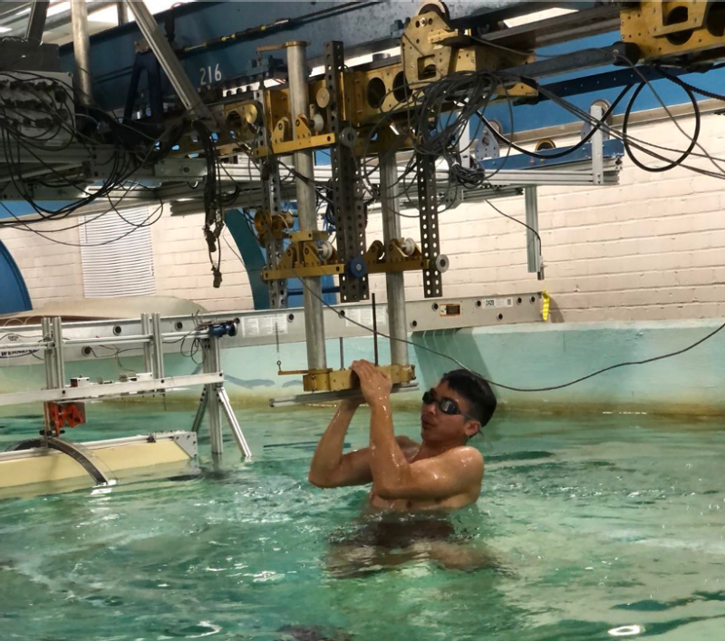This photo shows a young man with short dark hair and swim goggles in a pool. Above the pool is a complicate set of gears and pulleys and cords.d 