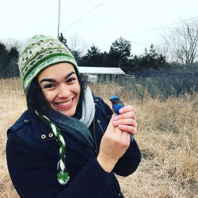 This photo shows a young woman with light brown skin, black hair, a green-and-white knit stocking cap, and a navy blue jacket. She is holding a bright blue bird in her hands and smiling at the camera. 