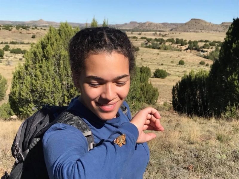 This photo shows a young woman with light brown skin and black hair, a medium blue long-sleeved shirt, a black backpack, and a small butterfly on her sleeve. In the background is a stretch of grasslands with occasional trees and mountains in the far distance. The woman is looking at the butterfly and smiling.